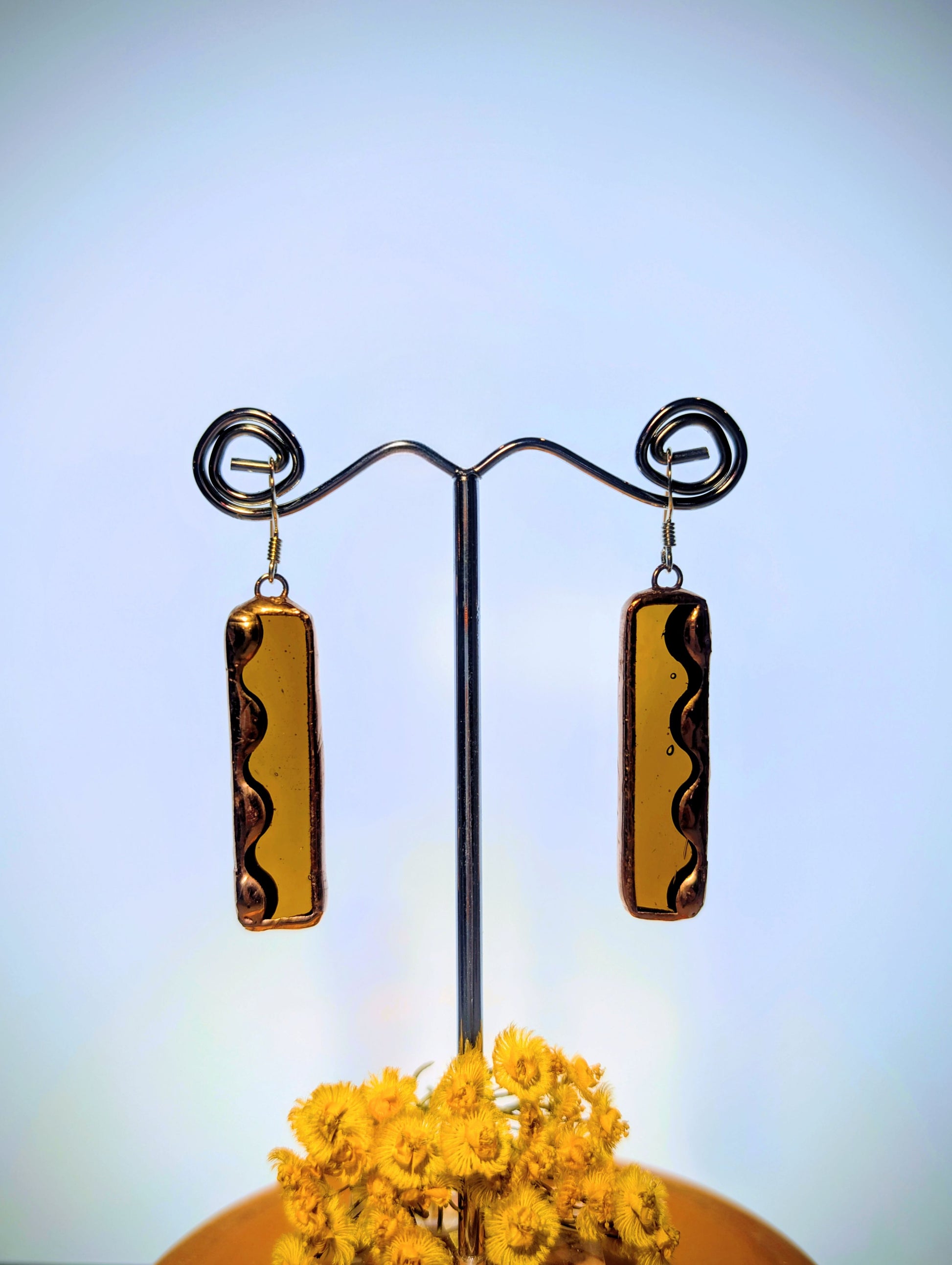 Pair of gold rectangular earrings on a stand with a blurred yellow flower in the foreground.