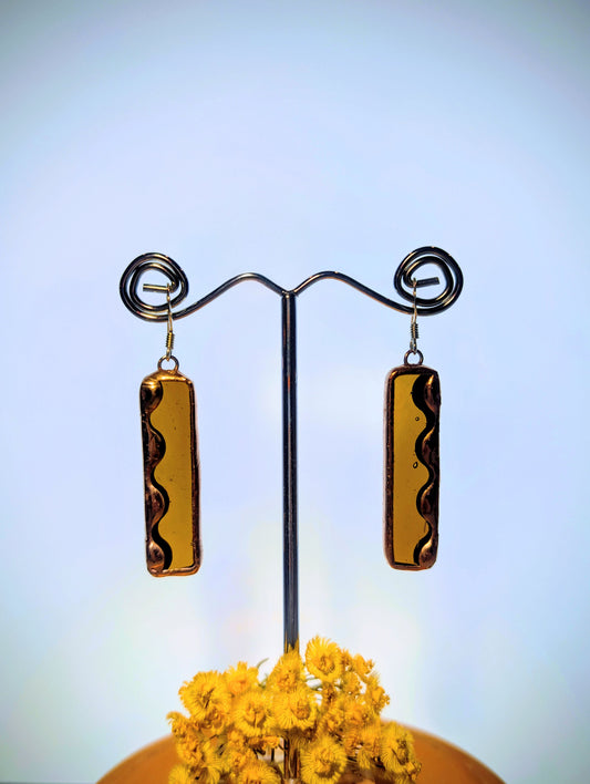 Pair of gold rectangular earrings on a stand with a blurred yellow flower in the foreground.