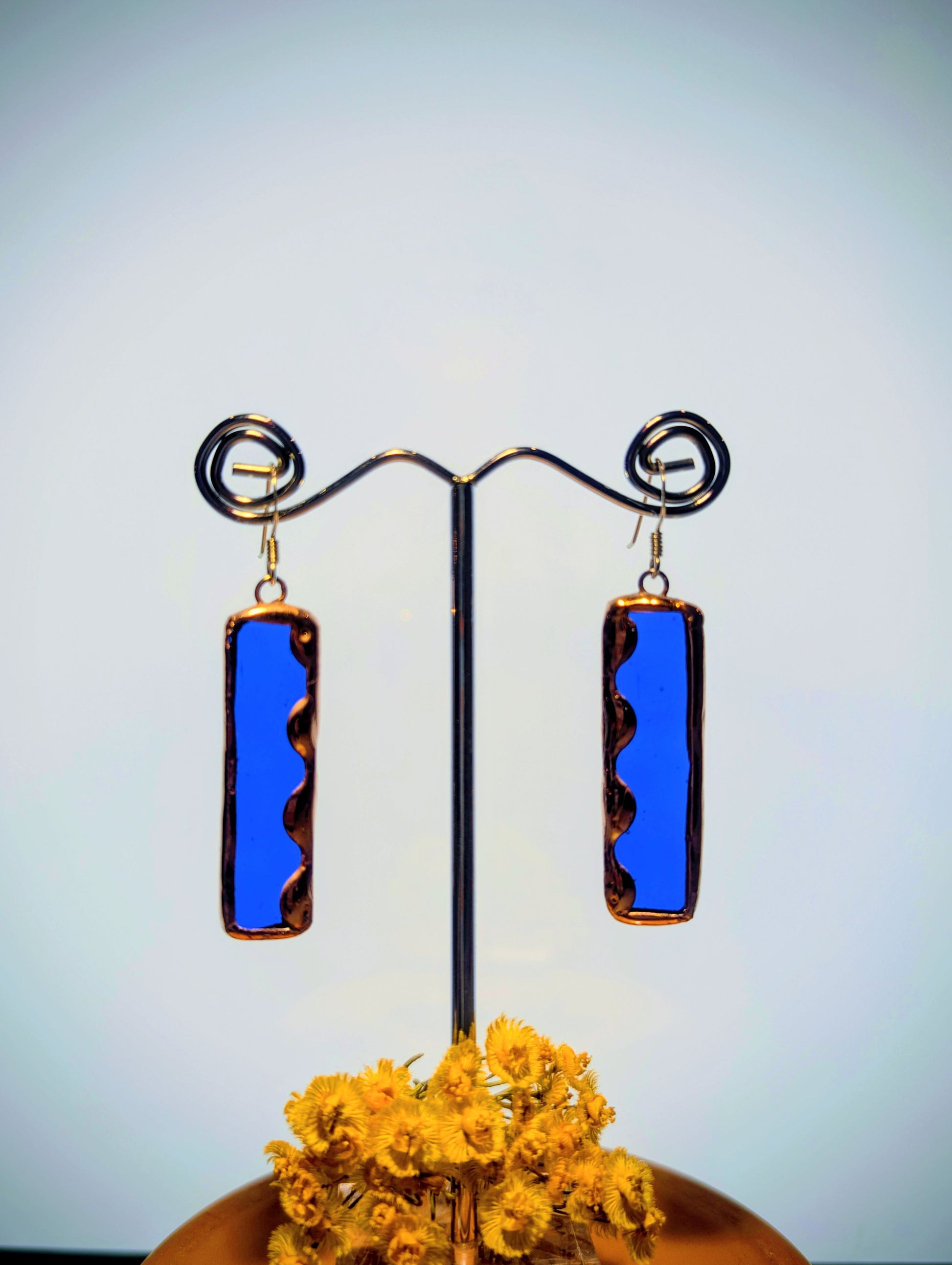 Pair of blue rectangular earrings on a stand with yellow flowers below.