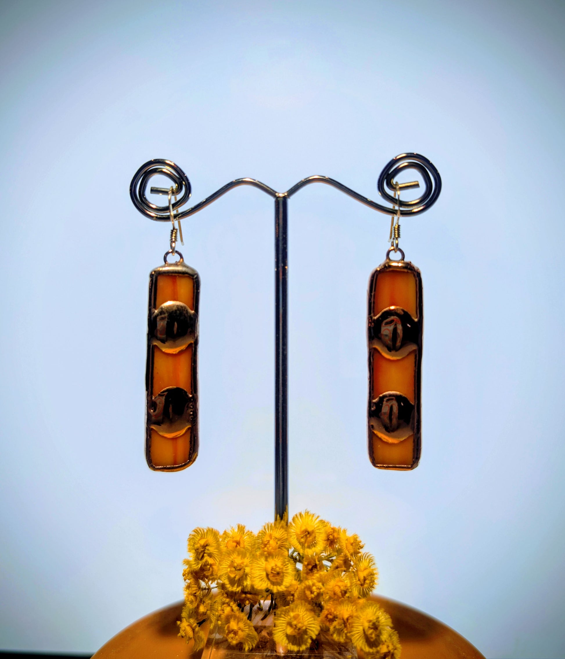 Amber-colored cylindrical earrings on a stand with yellow flowers below against a light blue background