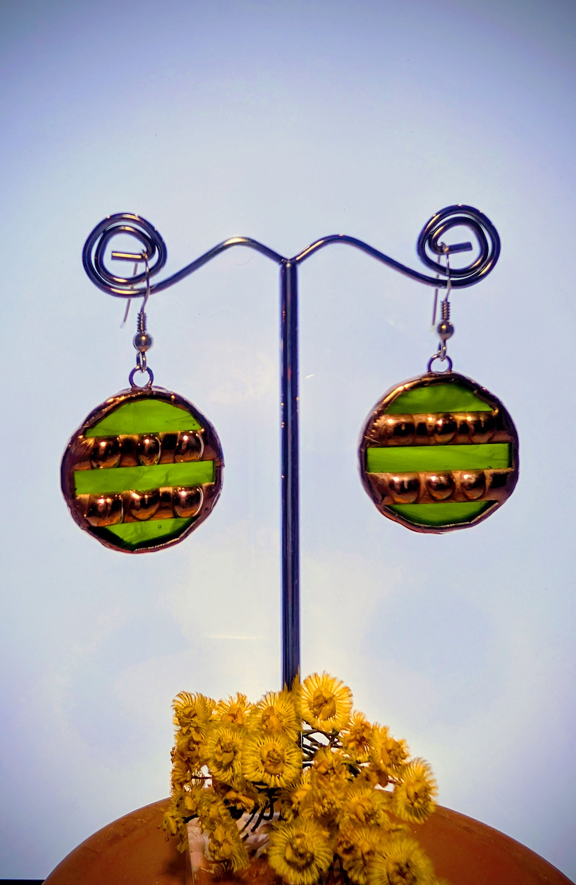 Pair of round earrings with green and gold design on a stand with yellow flowers below.