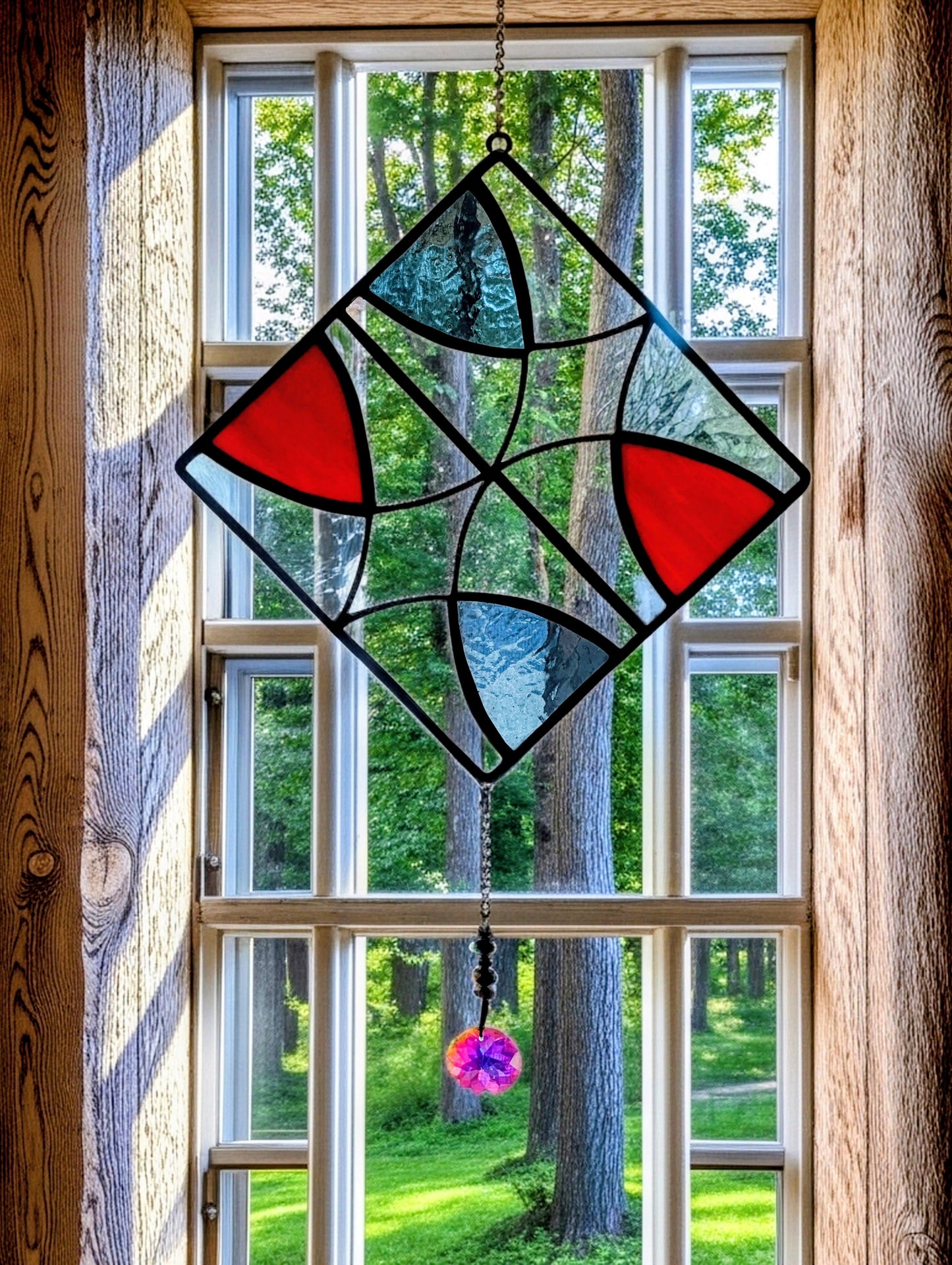 Stained glass window hanging with geometric design in front of a wooden window with a view of trees.